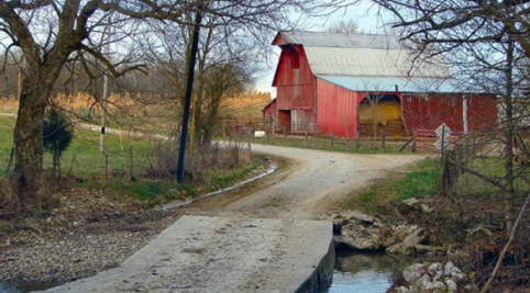 A photograph of a dirt road leading to a red barn with trees on each side